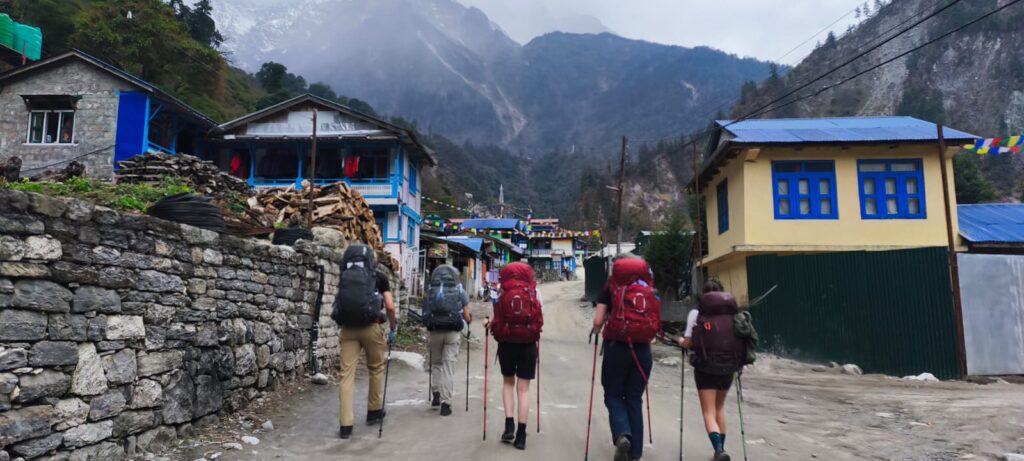 Trekkers hiking through a traditional mountain village in Nepal with colorful houses, prayer flags, and misty Himalayan mountains in the background.